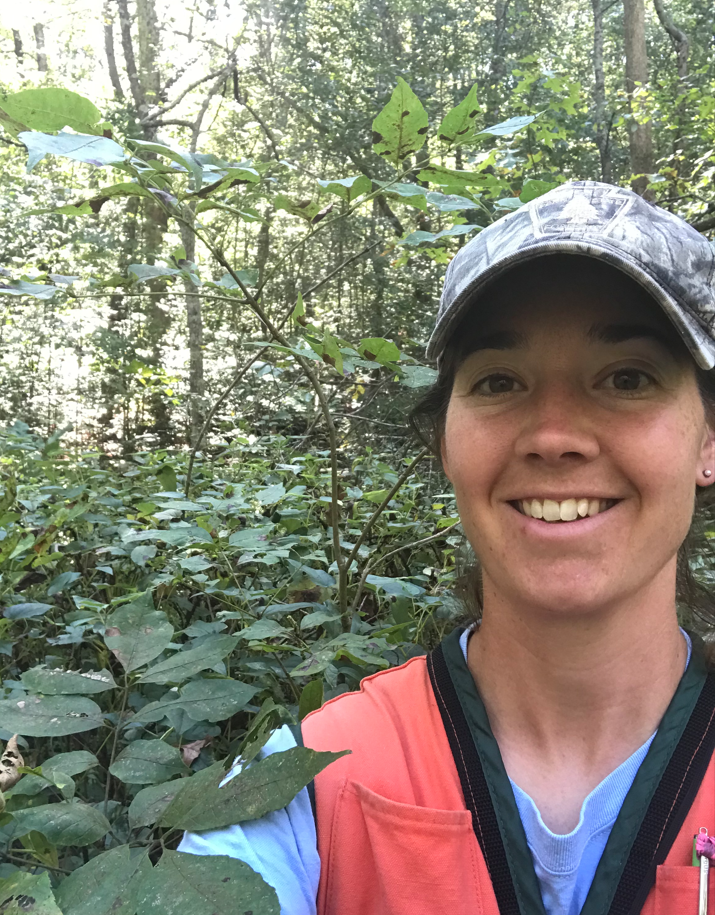 Jess smiling in the woods, wearing a camo baseball hat, orange vest, and blue shirt